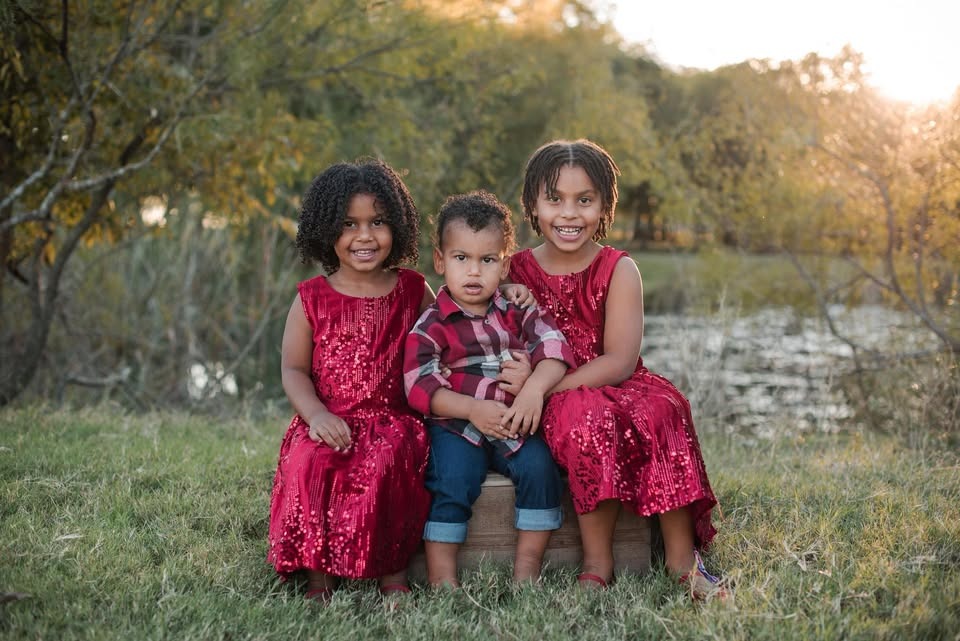 Asher held by his two sisters in red dresses beside a pond